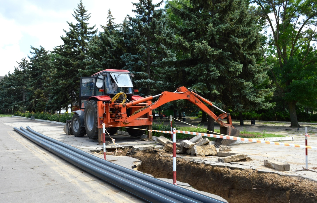 A red backhoe loader is parked near a construction trench on a road. Pipes are placed alongside the trench, and a safety barrier surrounds the area. Trees are in the background.