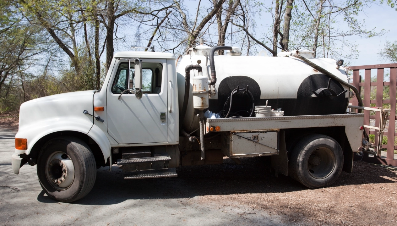 A white truck with a large cylindrical tank parked on a road surrounded by trees.