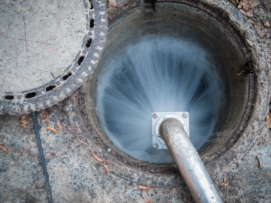 A metal pipe discharges water into an open manhole, creating a swirling motion. A manhole cover is partially removed and lies on the concrete ground.