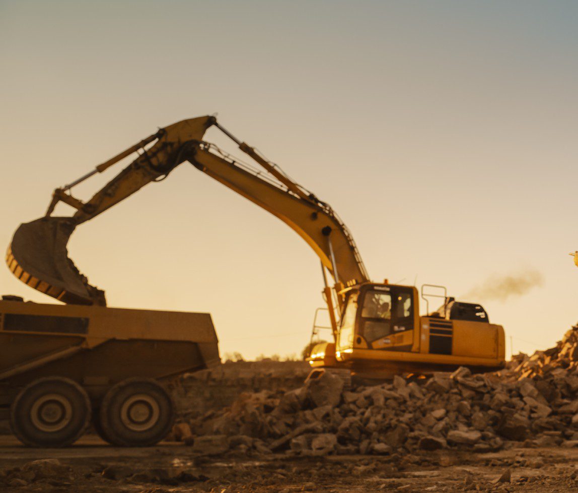 Excavator loading dirt into dump truck at sunset on a construction site.